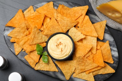 Delicious nachos and cheese sauce with basil on black table, flat lay
