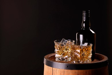 Photo of Whiskey with ice cubes in glasses and bottle on wooden barrel against dark background, space for text