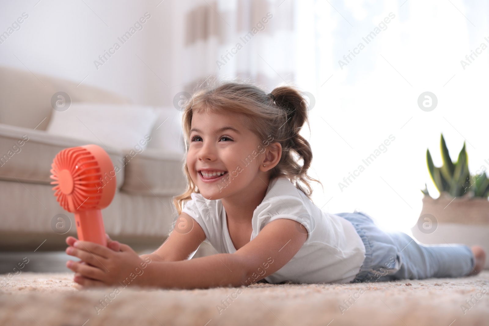 Photo of Little girl enjoying air flow from portable fan on floor in living room. Summer heat