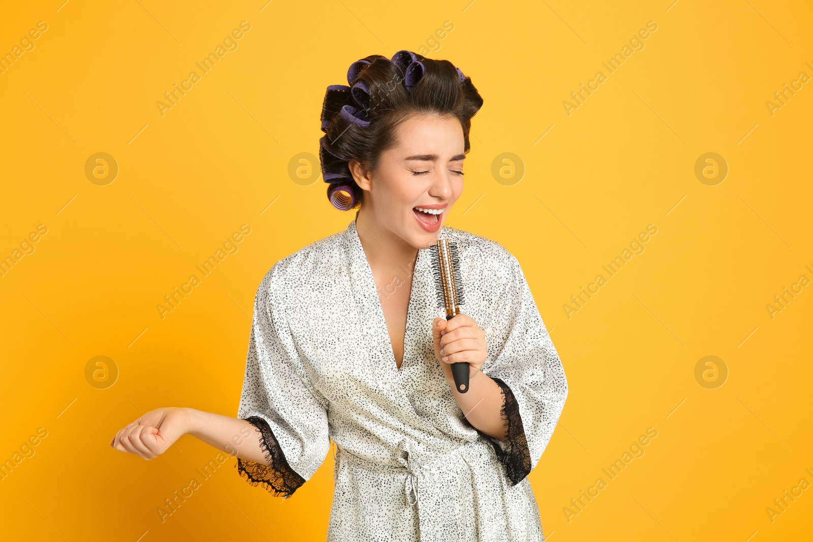 Photo of Happy young woman in silk bathrobe with hair curlers singing into hairbrush on orange background