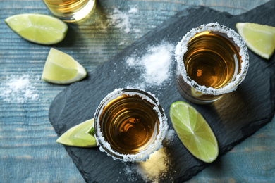 Photo of Mexican Tequila shots, lime slices and salt on blue wooden table, flat lay