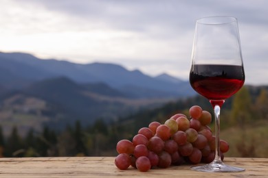 Glass of tasty red wine and grapes on wooden table against mountain landscape. Space for text