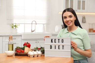 Photo of Happy woman holding lightbox with words Keto Diet near different products in kitchen