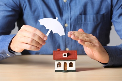 Man covering house model with umbrella cutout at table, closeup. Home insurance