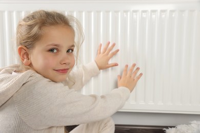 Photo of Little girl warming hands near heating radiator indoors