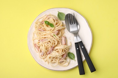 Photo of Plate of tasty pasta Carbonara with basil leaves, fork and spoon on yellow background, top view