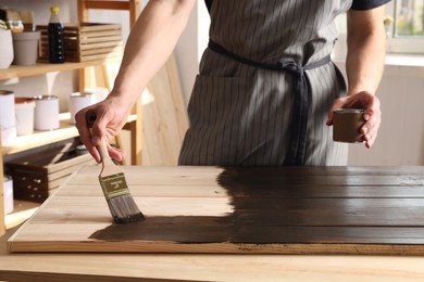 Photo of Man with brush and can applying wood stain onto wooden surface indoors, closeup
