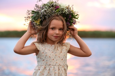 Photo of Cute little girl wearing wreath madebeautiful flowers near river at sunset