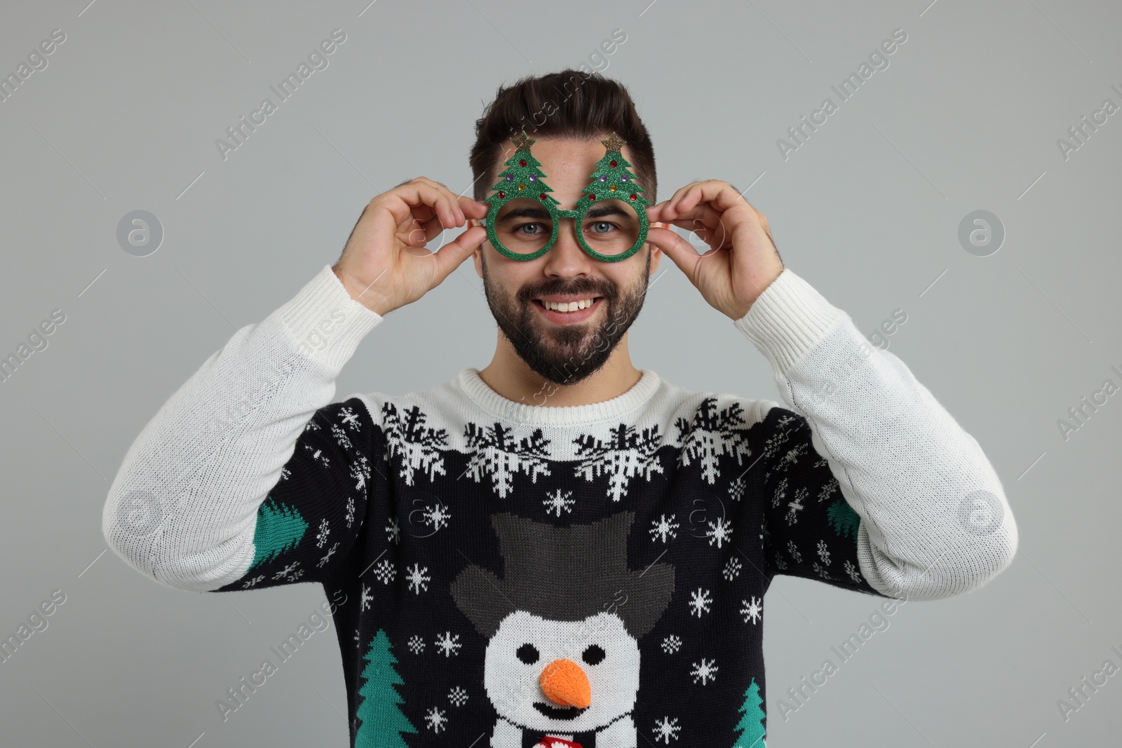 Photo of Happy young man in Christmas sweater and funny glasses on grey background
