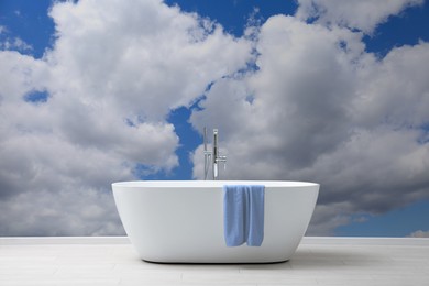 Image of Blue sky with clouds as wallpaper pattern in room. Ceramic bathtub with towel near wall