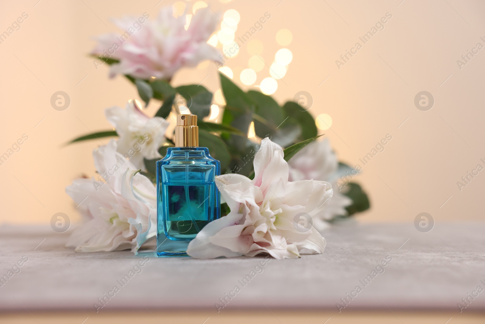 Photo of Bottle of perfume and beautiful lily flowers on table against beige background with blurred lights, closeup