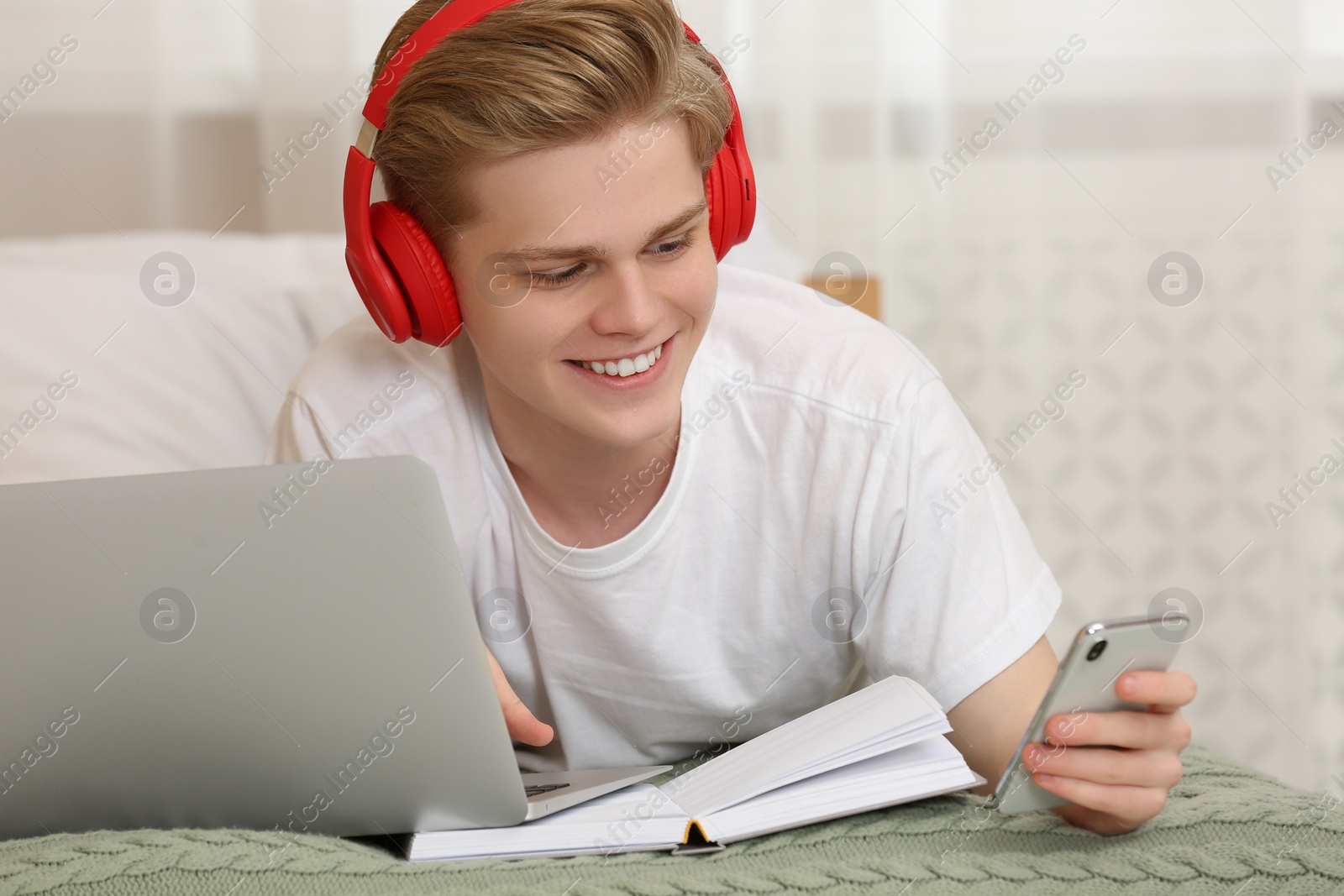 Photo of Teenage boy with smartphone and headphones using laptop at home