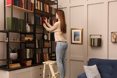 Photo of Young woman choosing book on shelf in home library