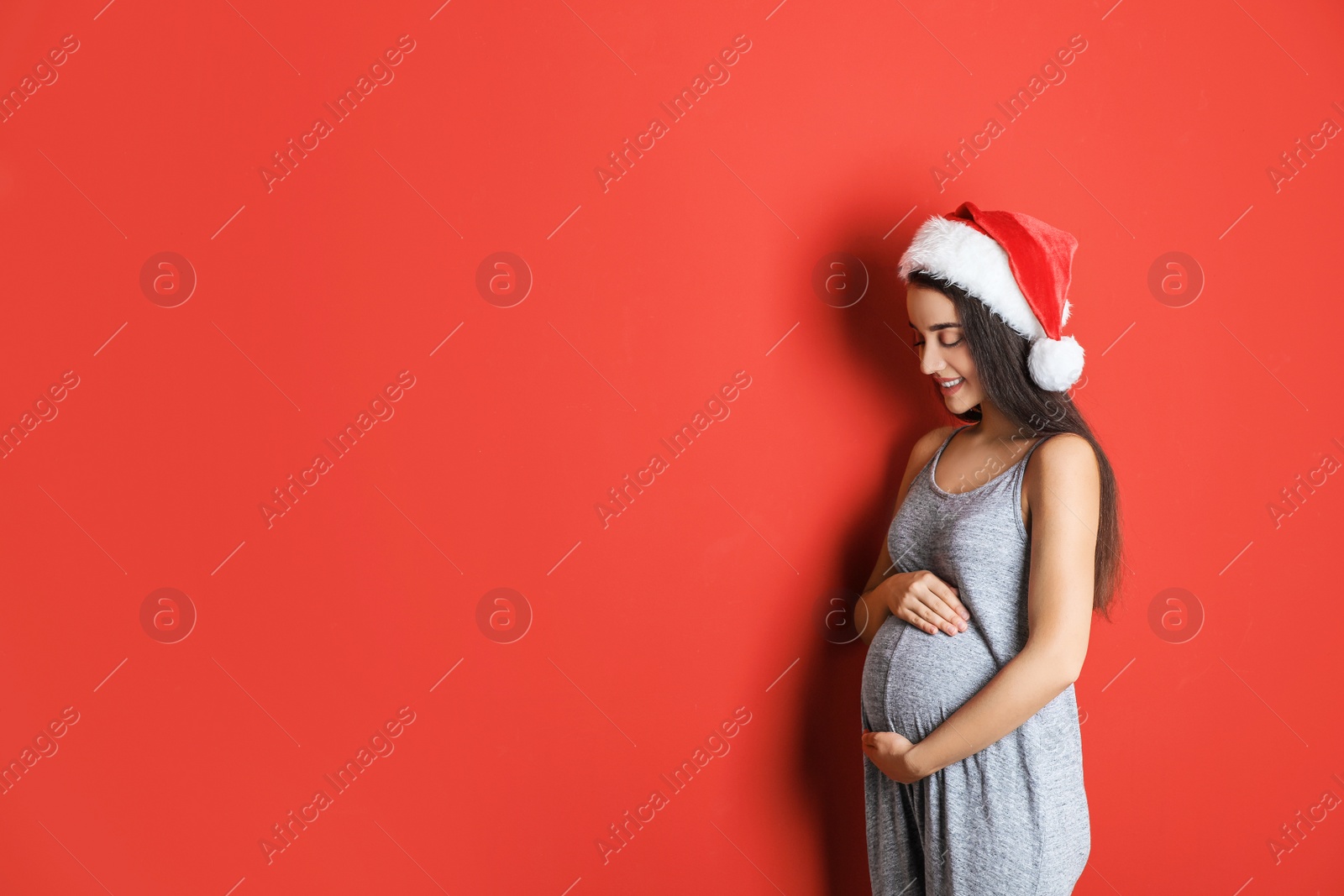 Photo of Happy pregnant woman wearing Santa hat for Christmas party on red background, space for text. Expecting baby