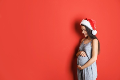 Happy pregnant woman wearing Santa hat for Christmas party on red background, space for text. Expecting baby