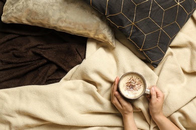 Photo of Woman holding cup of coffee while lying on bed with pillows and warm plaid, top view