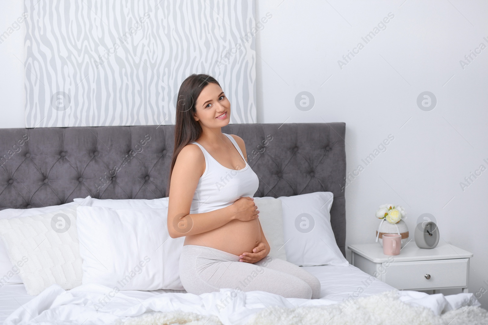 Photo of Young beautiful pregnant woman sitting on bed and touching her belly at home