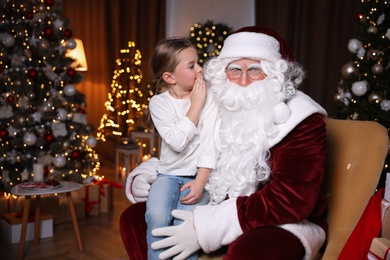 Photo of Little girl whispering in Santa Claus' ear near Christmas tree indoors