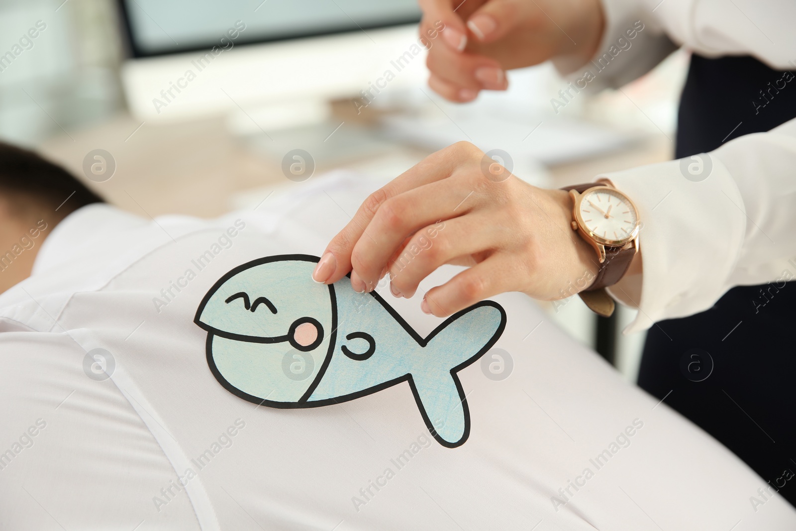 Photo of Young woman sticking paper fish to colleague's back in office, closeup. Funny joke