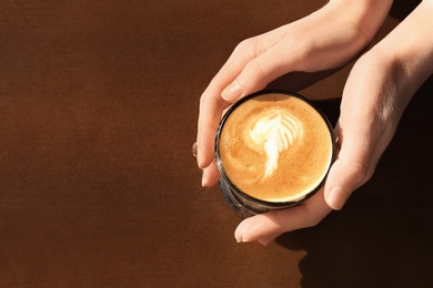 Woman holding cup of aromatic coffee with foam at wooden table, top view