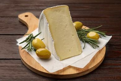 Photo of Board with piece of tasty camembert cheese, olives and rosemary on wooden table, closeup