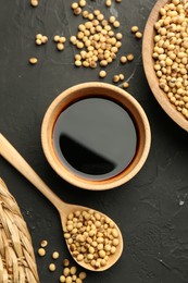 Photo of Soy sauce in bowl and soybeans on black textured table, flat lay