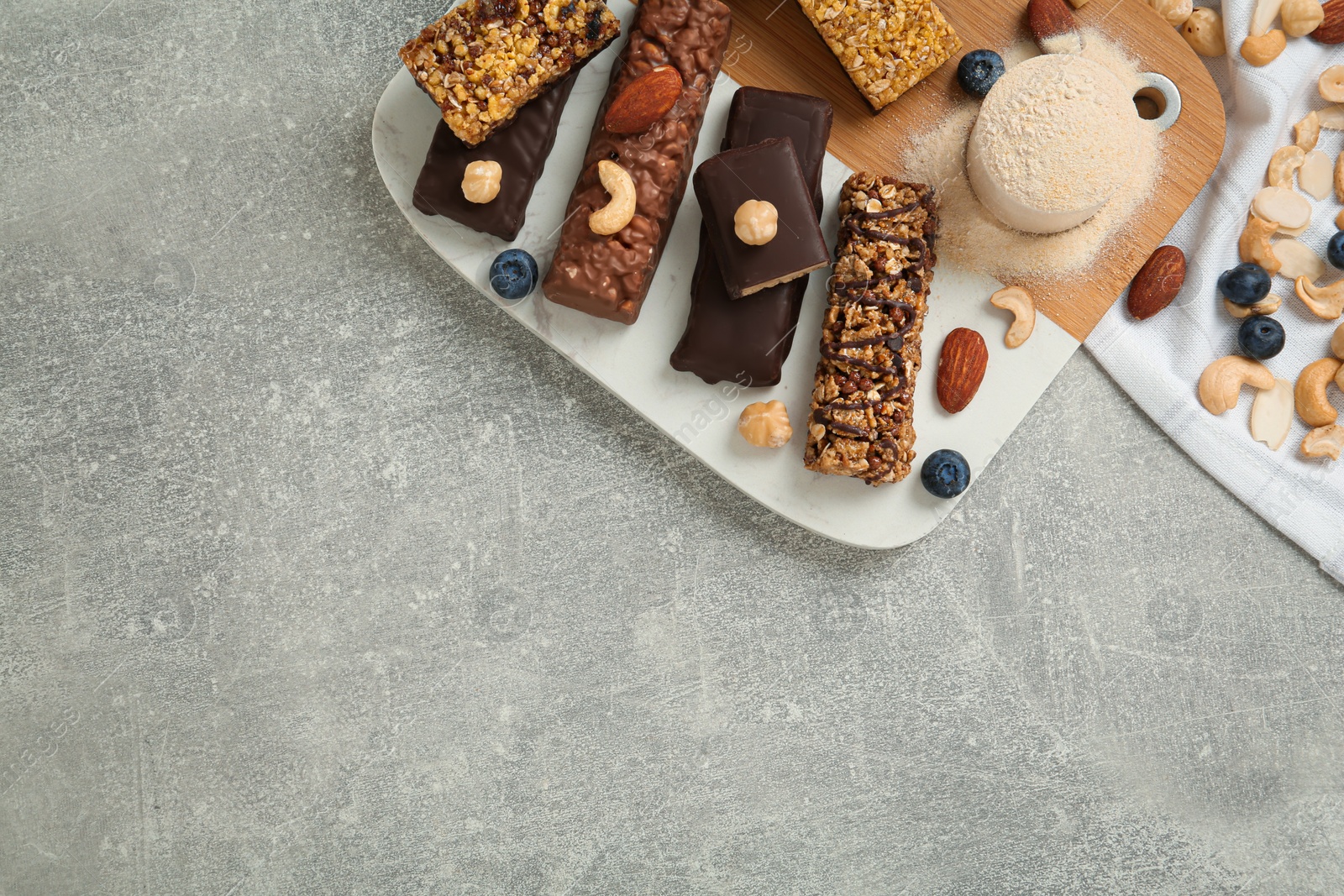 Photo of Different energy bars, nuts, blueberries and protein powder on grey table, flat lay. Space for text