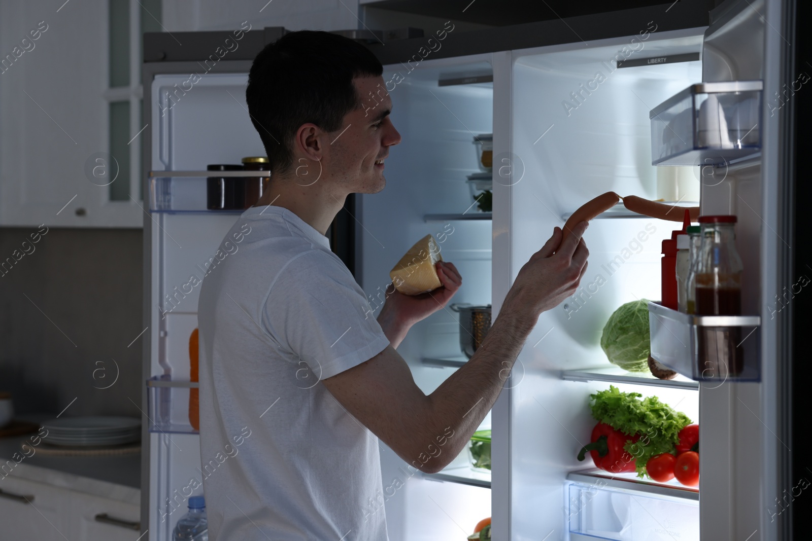 Photo of Man with cheese taking sausages out of refrigerator in kitchen at night