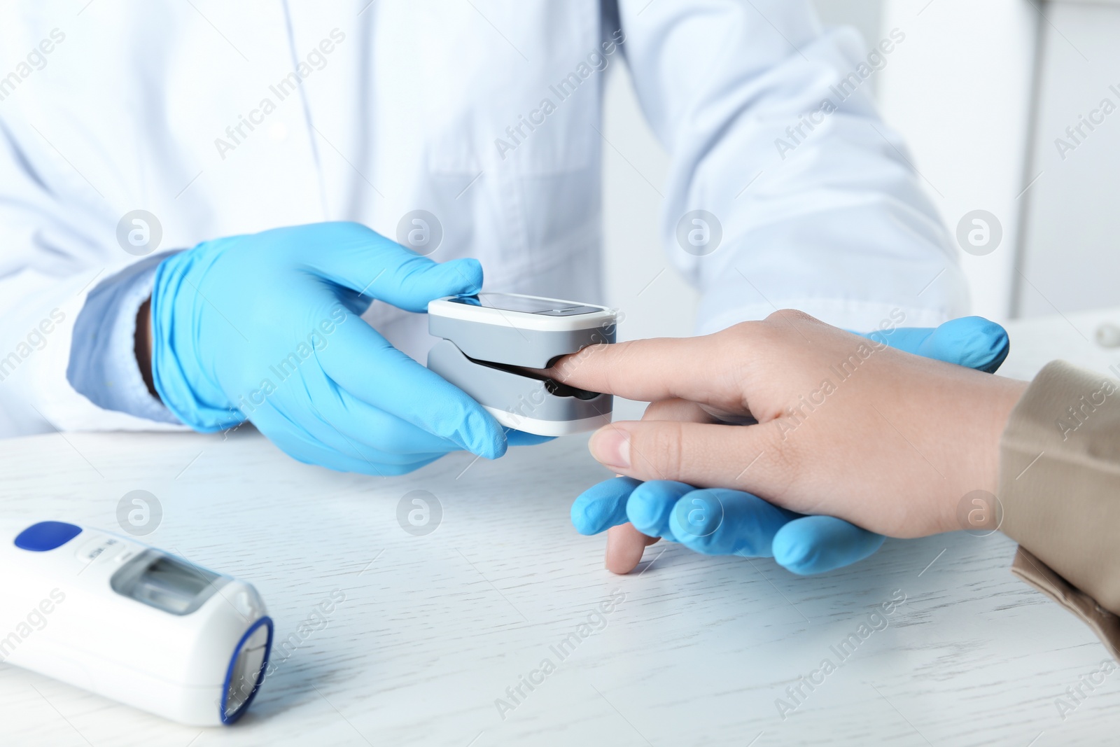 Photo of Doctor examining patient with fingertip pulse oximeter at white wooden table, closeup