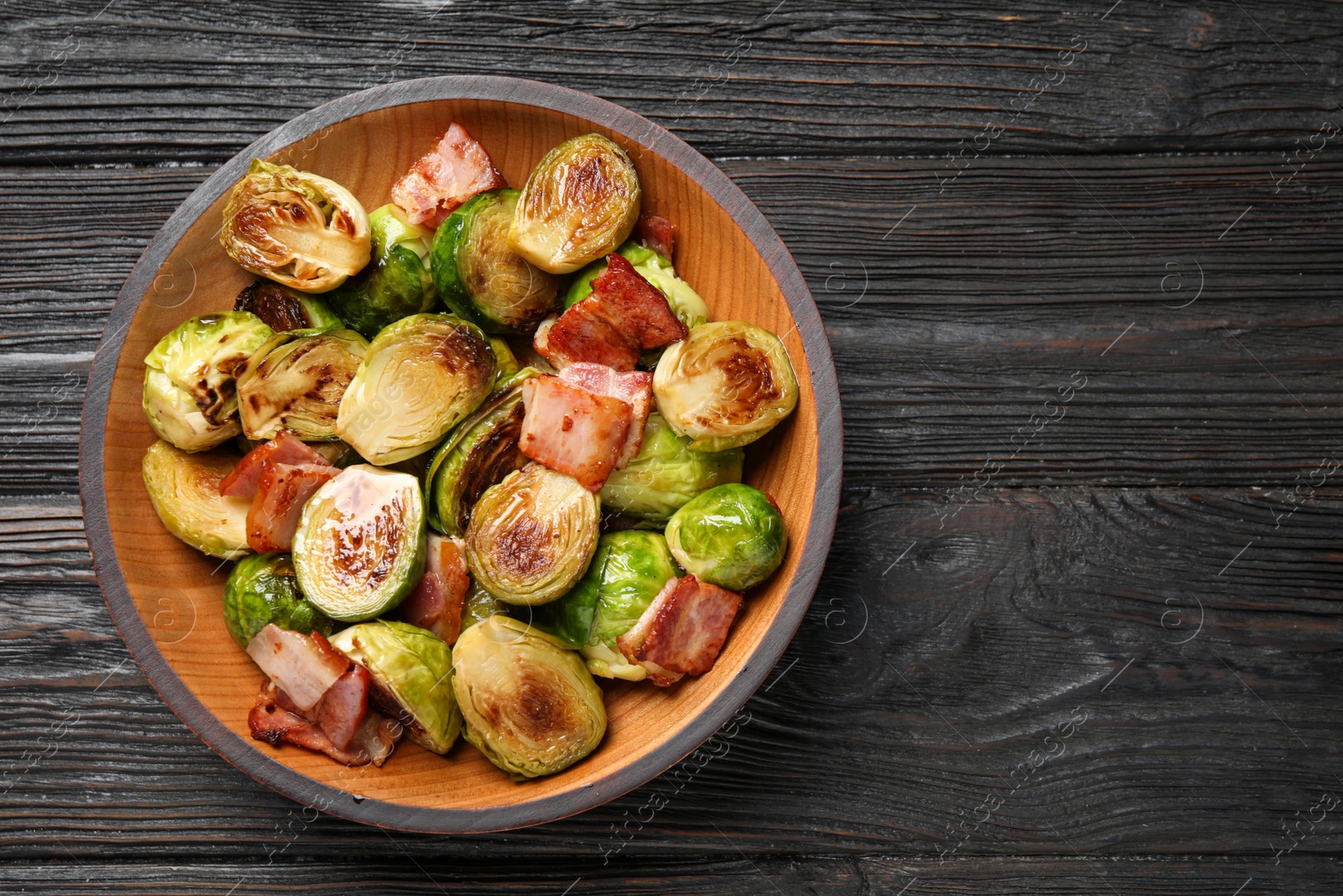 Image of Delicious fried Brussels sprouts with bacon in bowl on wooden table, top view. Space for text