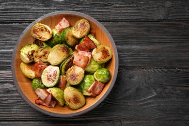 Delicious fried Brussels sprouts with bacon in bowl on wooden table, top view. Space for text