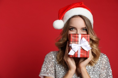 Happy young woman wearing Santa hat with Christmas gift on red background, space for text