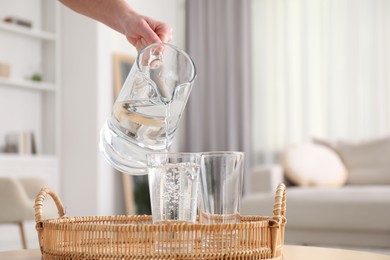 Photo of Woman pouring fresh water from jug into glass at table indoors, closeup. Space for text