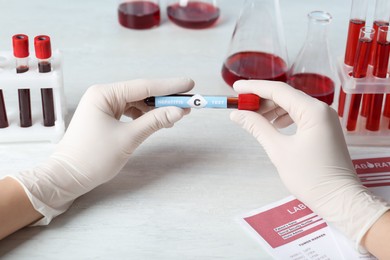 Scientist holding tube with blood sample and label Hepatitis C test at white wooden table, closeup