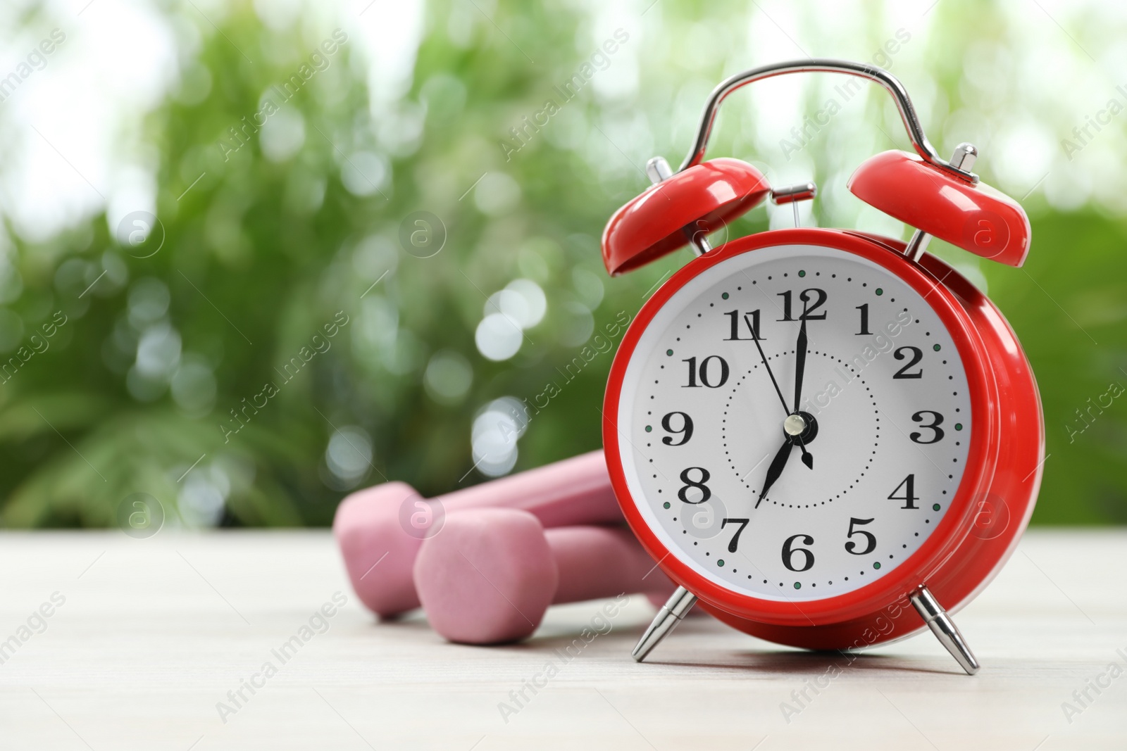 Photo of Alarm clock and dumbbells on white wooden table outdoors, space for text. Morning exercise