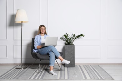 Young woman with laptop in armchair at home, space for text. Interior design