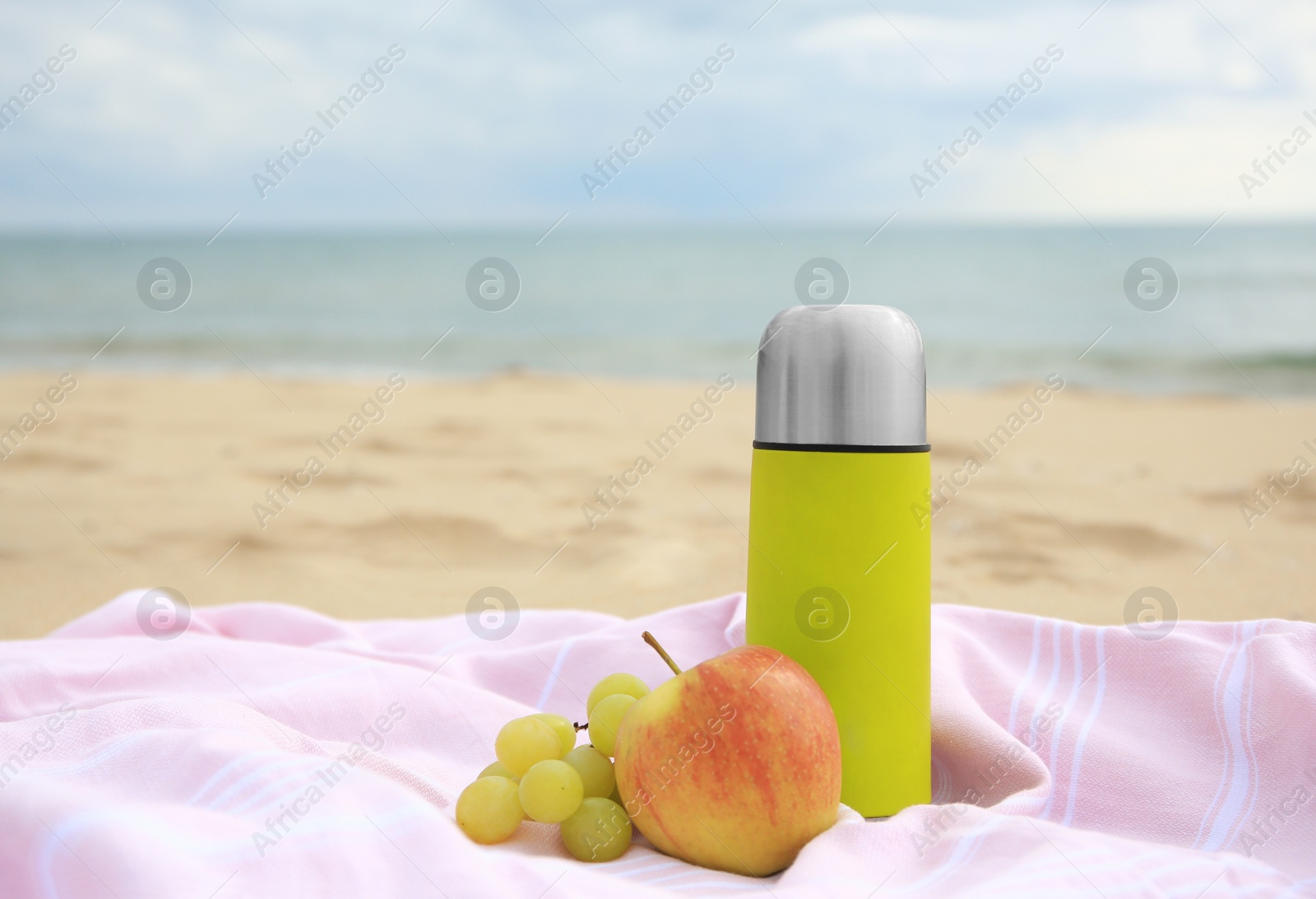 Photo of Metallic thermos with hot drink, fruits and plaid on sandy beach near sea