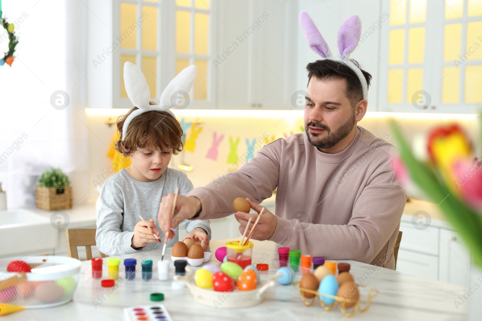 Photo of Easter celebration. Father with his little son painting eggs at white marble table in kitchen