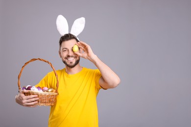 Photo of Happy man in bunny ears headband holding wicker basket with painted Easter eggs on grey background. Space for text