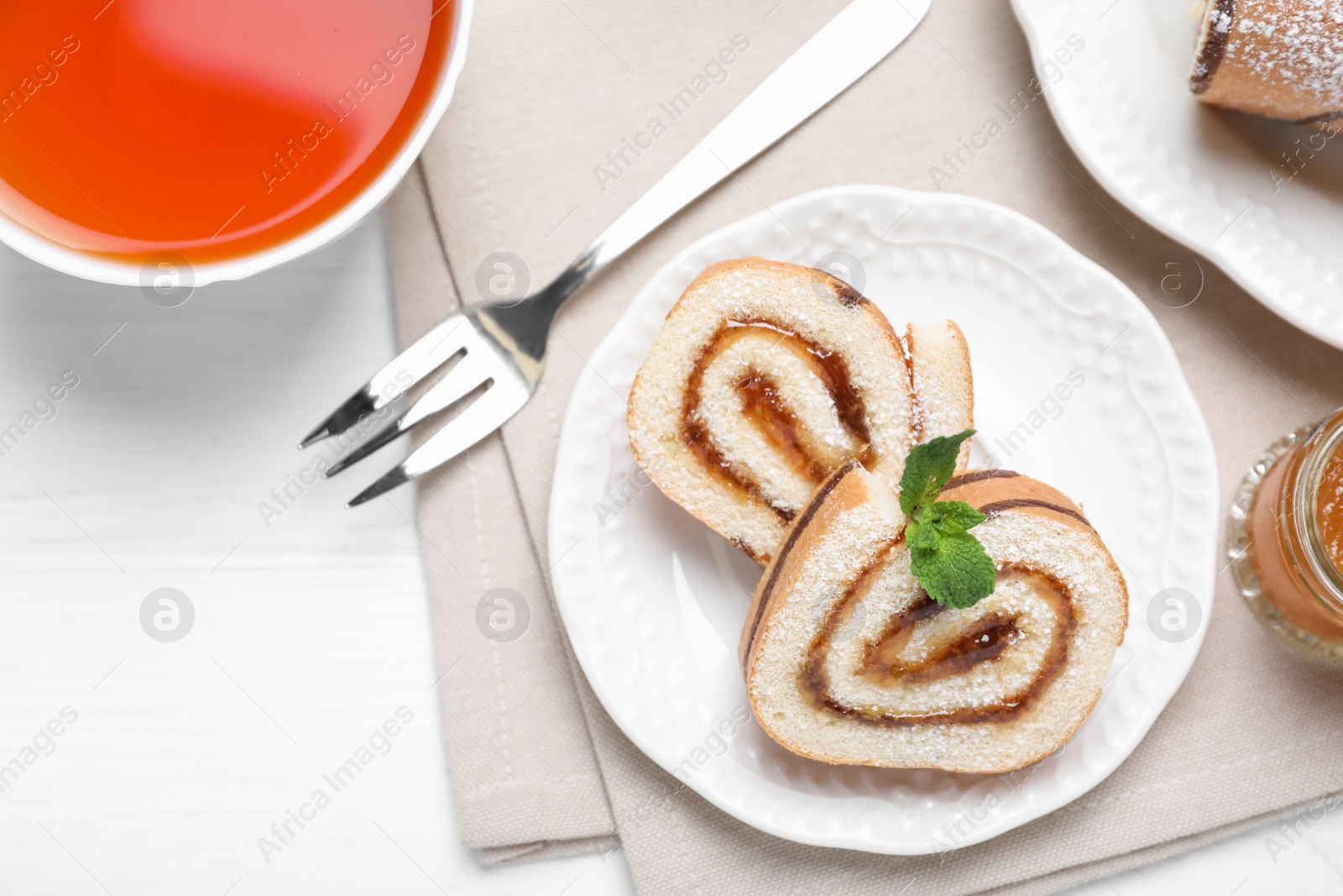 Photo of Tasty cake roll with jam and cup of tea on white wooden table, flat lay