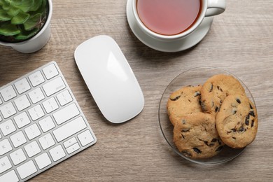 Photo of Chocolate chip cookies, tea and keyboard on wooden table at workplace, flat lay