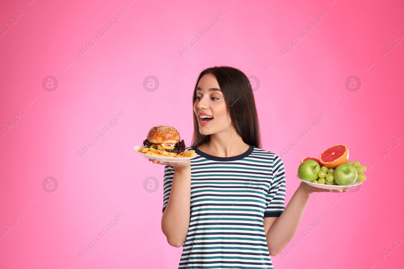 Photo of Woman choosing between fruits and burger with French fries on pink background