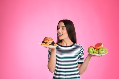 Photo of Woman choosing between fruits and burger with French fries on pink background