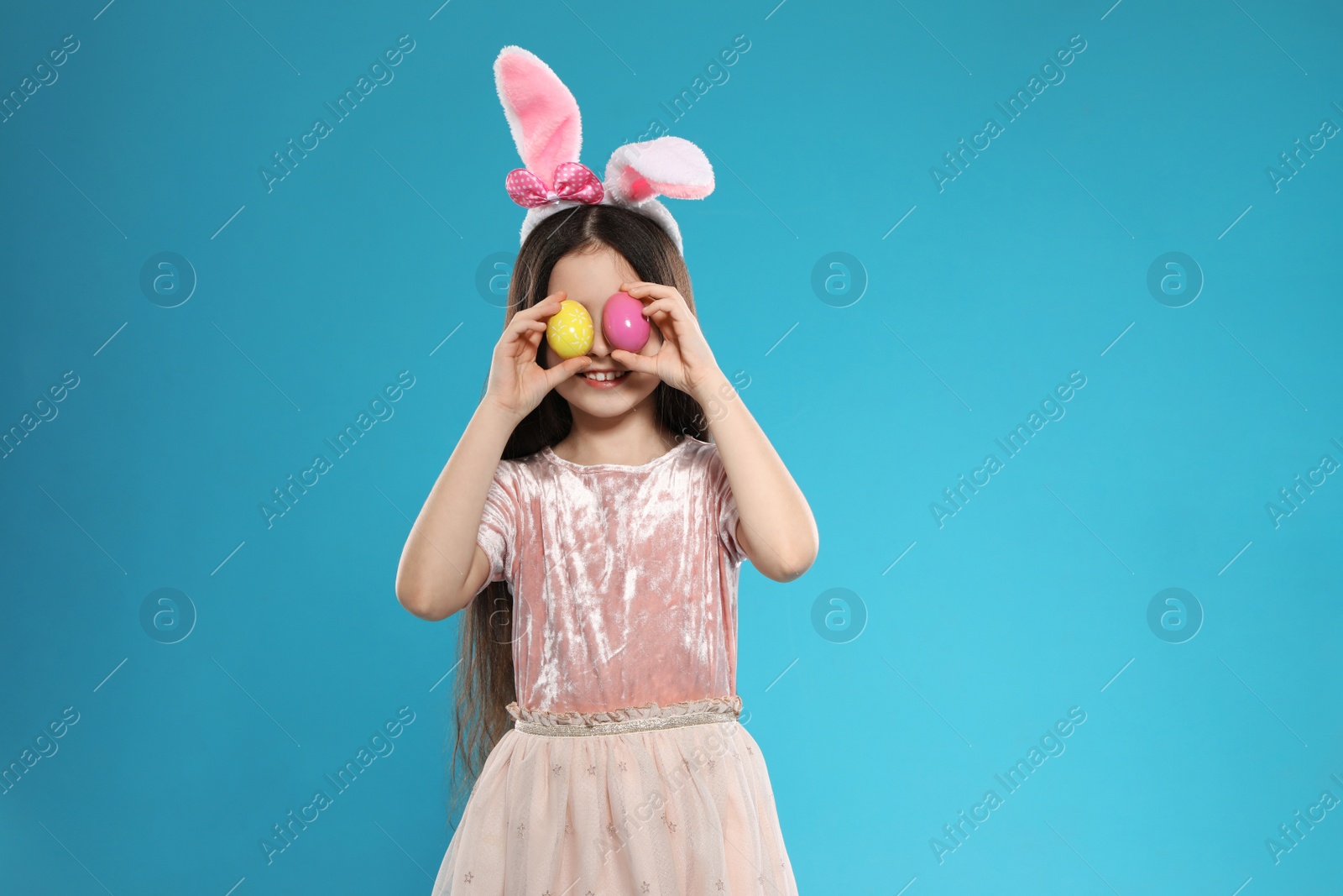 Photo of Little girl in bunny ears headband holding Easter eggs near eyes on color background