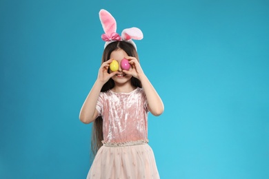 Little girl in bunny ears headband holding Easter eggs near eyes on color background