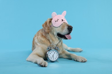 Photo of Cute Labrador Retriever with sleep mask and alarm clock resting on light blue background