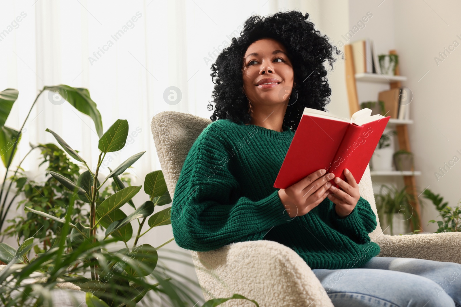 Photo of Relaxing atmosphere. Happy woman with book on armchair near beautiful houseplants in room
