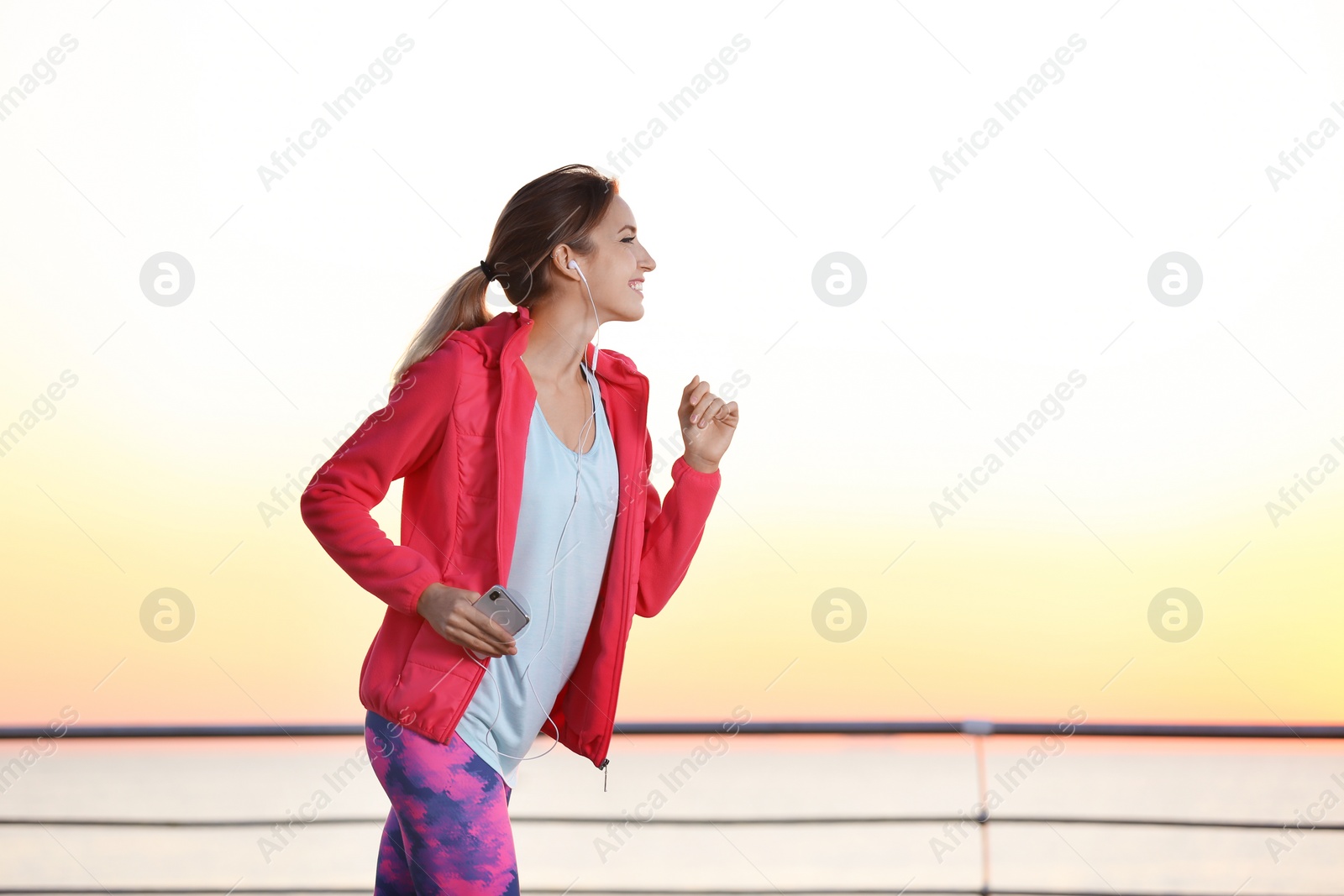 Photo of Young woman listening to music and running on pier in morning