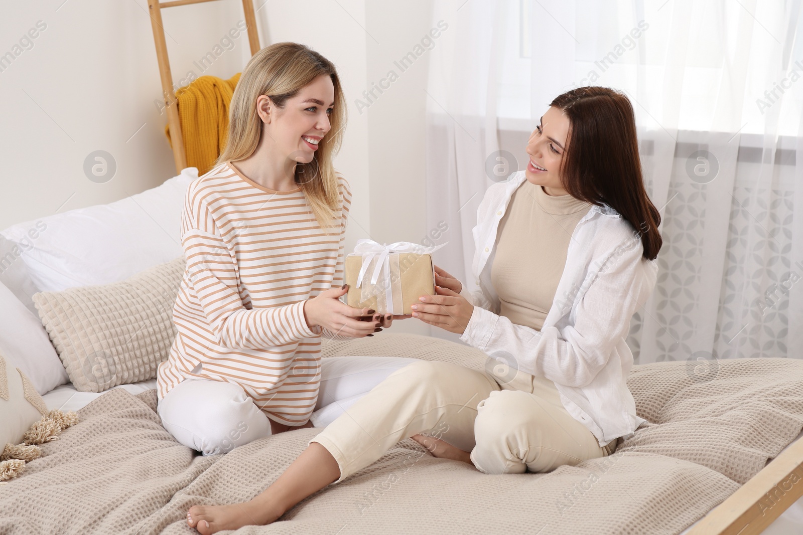 Photo of Smiling young woman presenting gift to her friend on bed at home