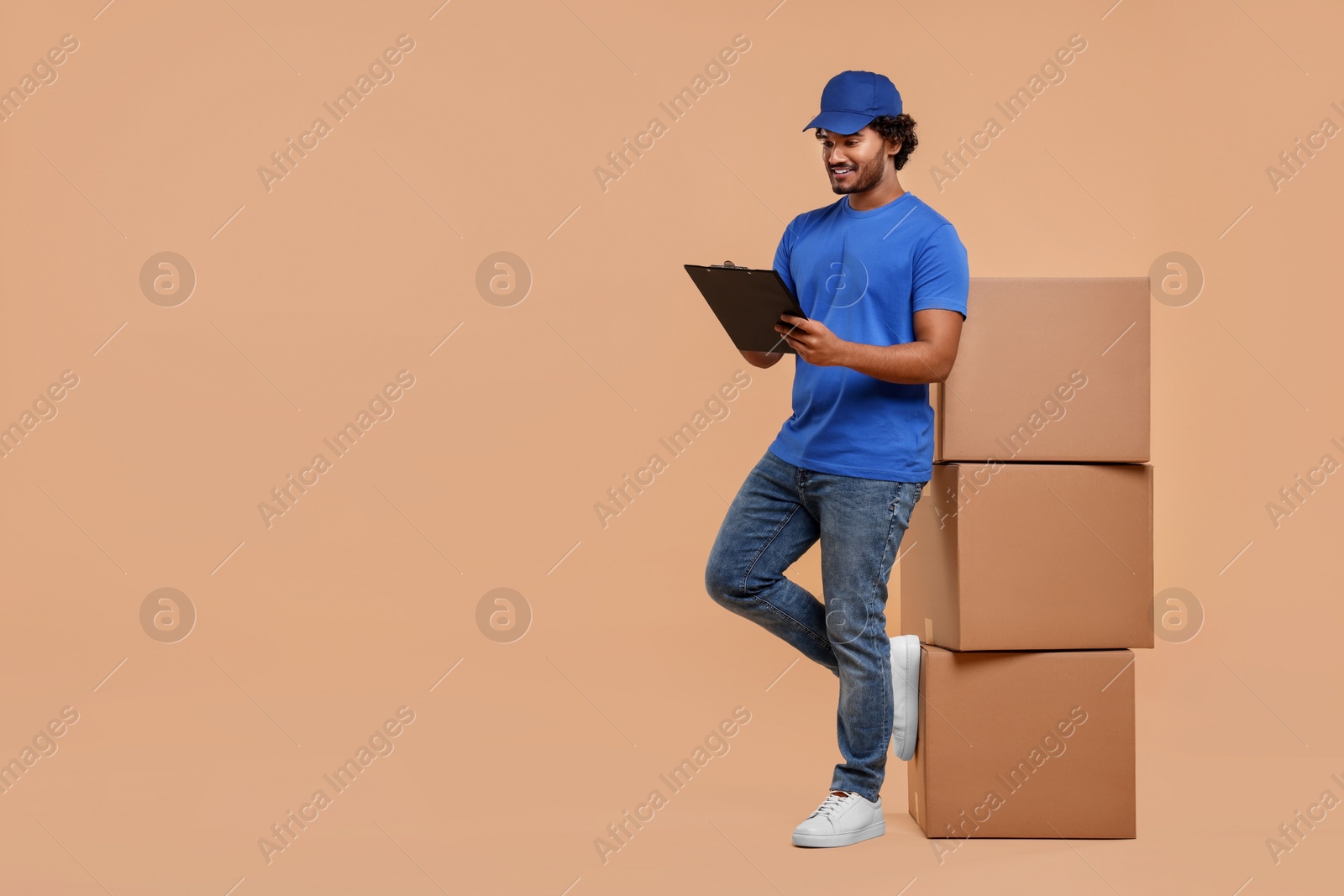 Photo of Happy courier with clipboard and stack of parcels on light brown background, space for text